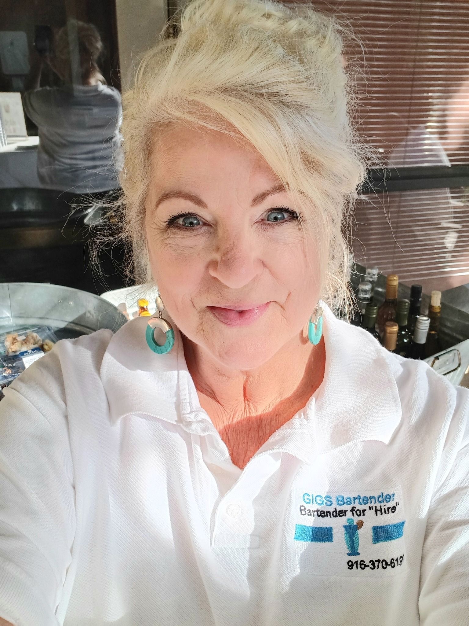 Woman with blonde hair wearing a white polo shirt smiles. Turquoise earrings, kitchen setting.