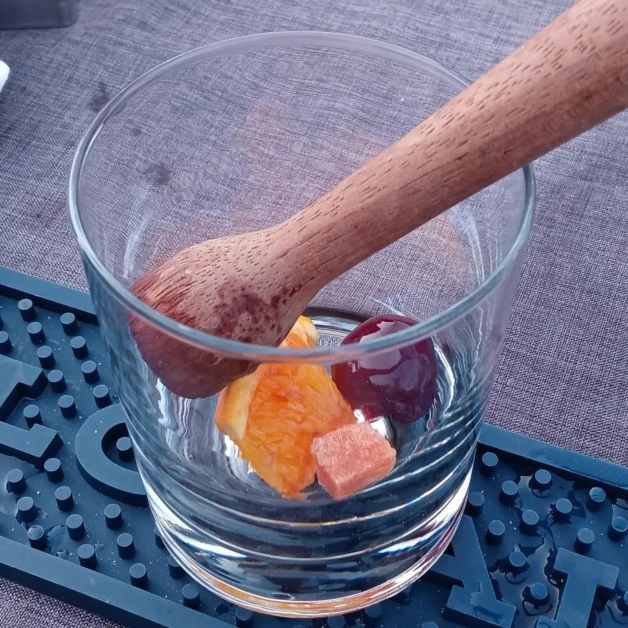 Cocktail glass with fruit, sugar cube, and muddler, resting on a black bar mat.