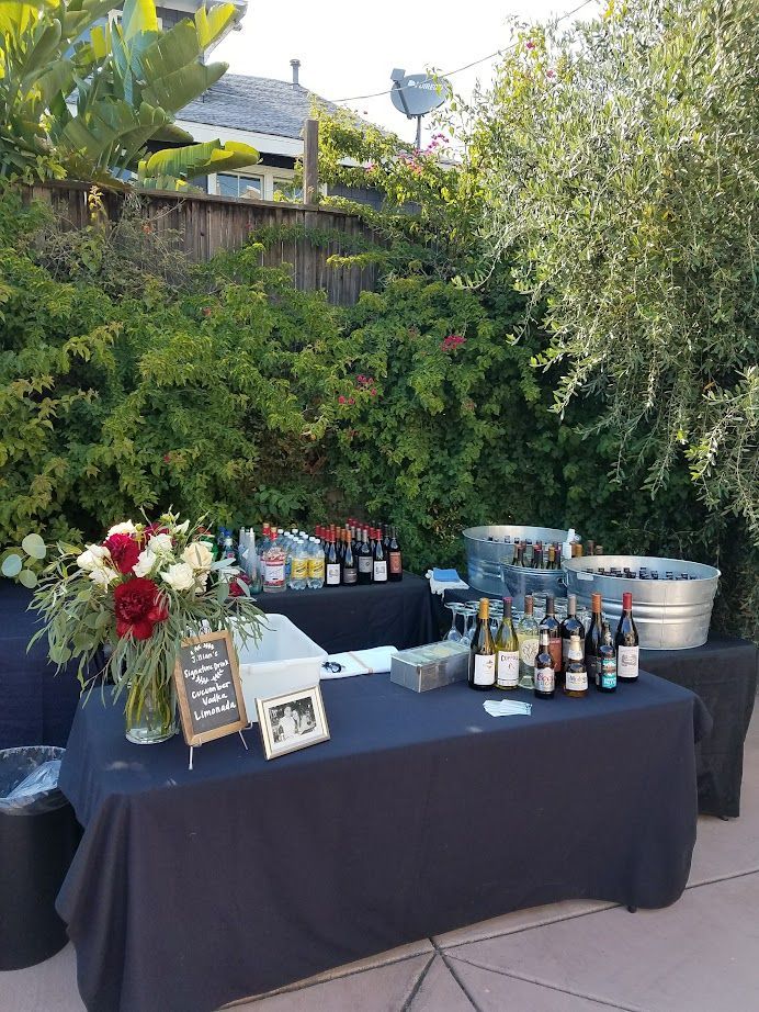 Outdoor bar with tables covered in black cloths, stocked with drinks, flowers, and decorations.