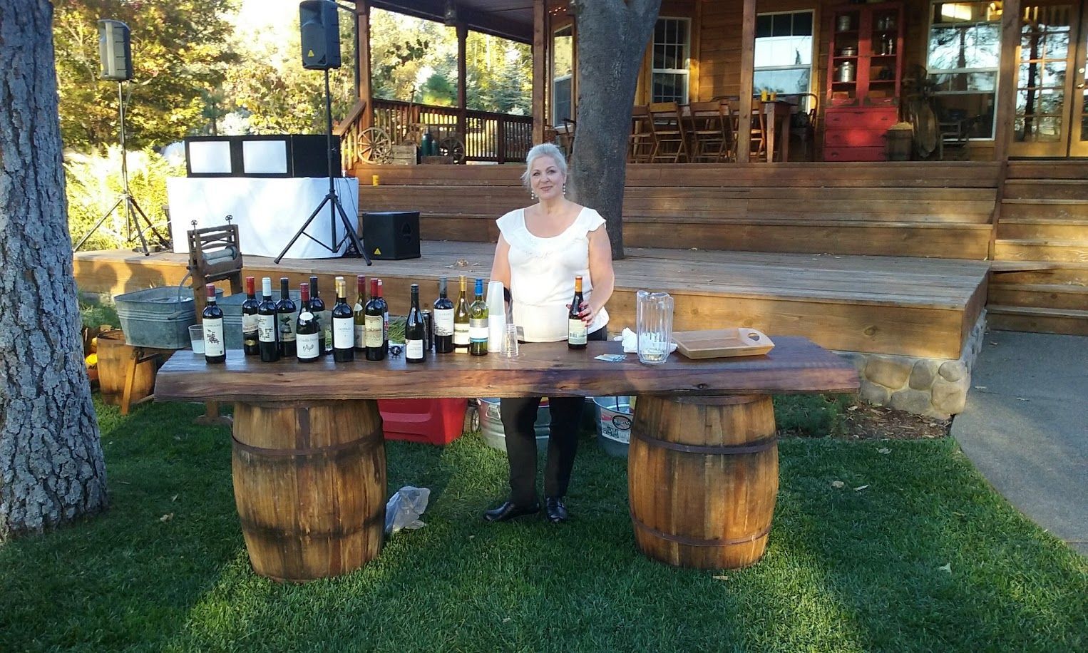 Woman at a wine tasting table, supported by barrels, outdoors near a wooden building.