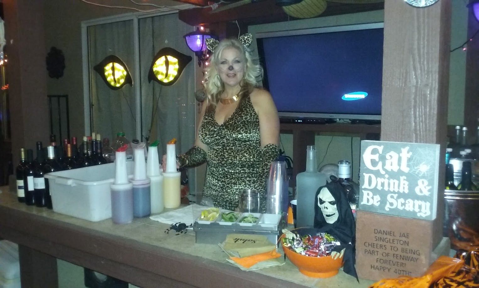 Woman in leopard costume at a Halloween bar. She stands behind a bar with drinks and decorations.