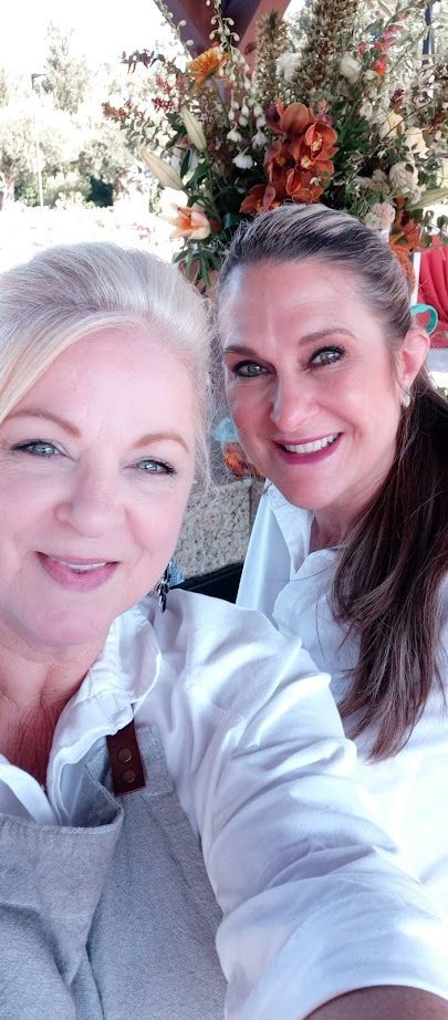 Two women smiling for a selfie, surrounded by fall flowers. One has blonde hair, the other brunette.