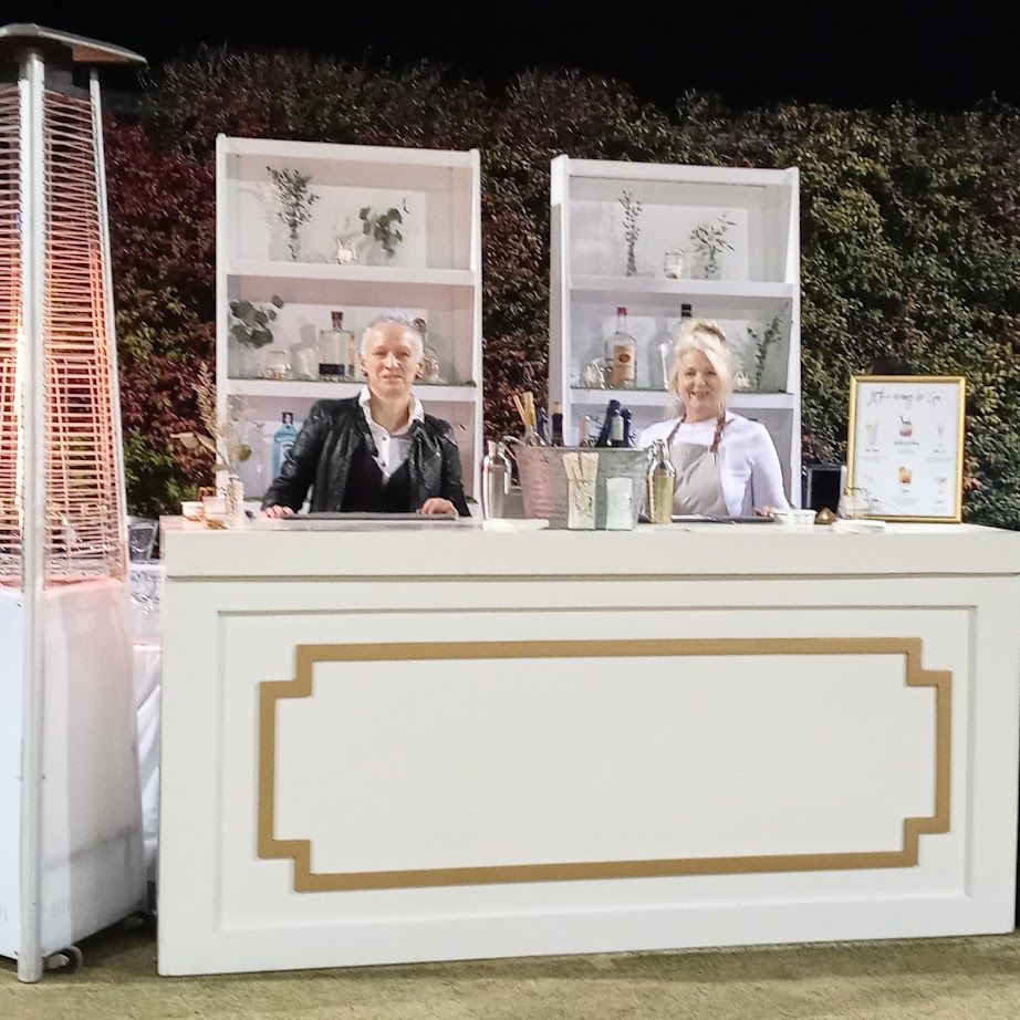 Two bartenders behind a white and gold bar, outdoor setting with a heater.