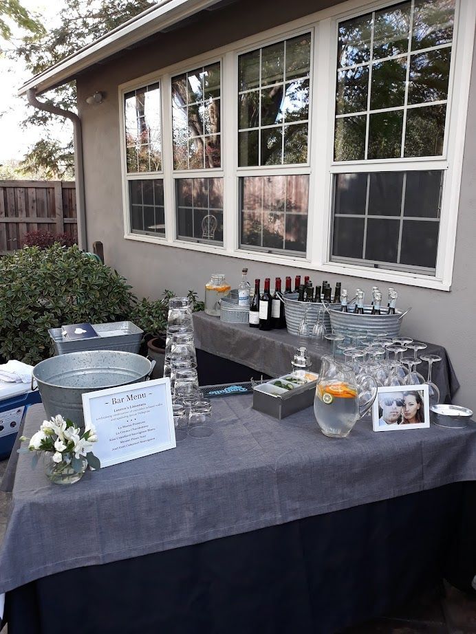 Outdoor bar with drinks, glasses, and a photo, set up against a building with large windows.