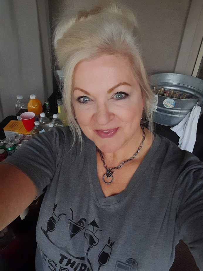Blonde woman in gray shirt smiles, indoors. Updo hairstyle, dark necklace. Bottles and tub visible in background.