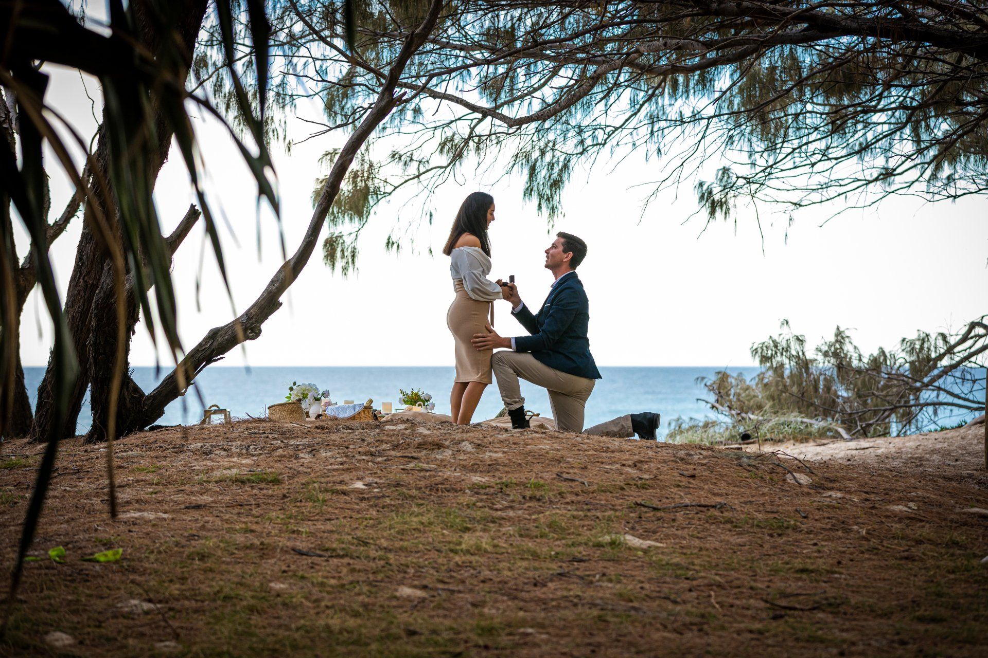 Engagement Shoot - a man is kneeling down to propose to a woman in front of the ocean .