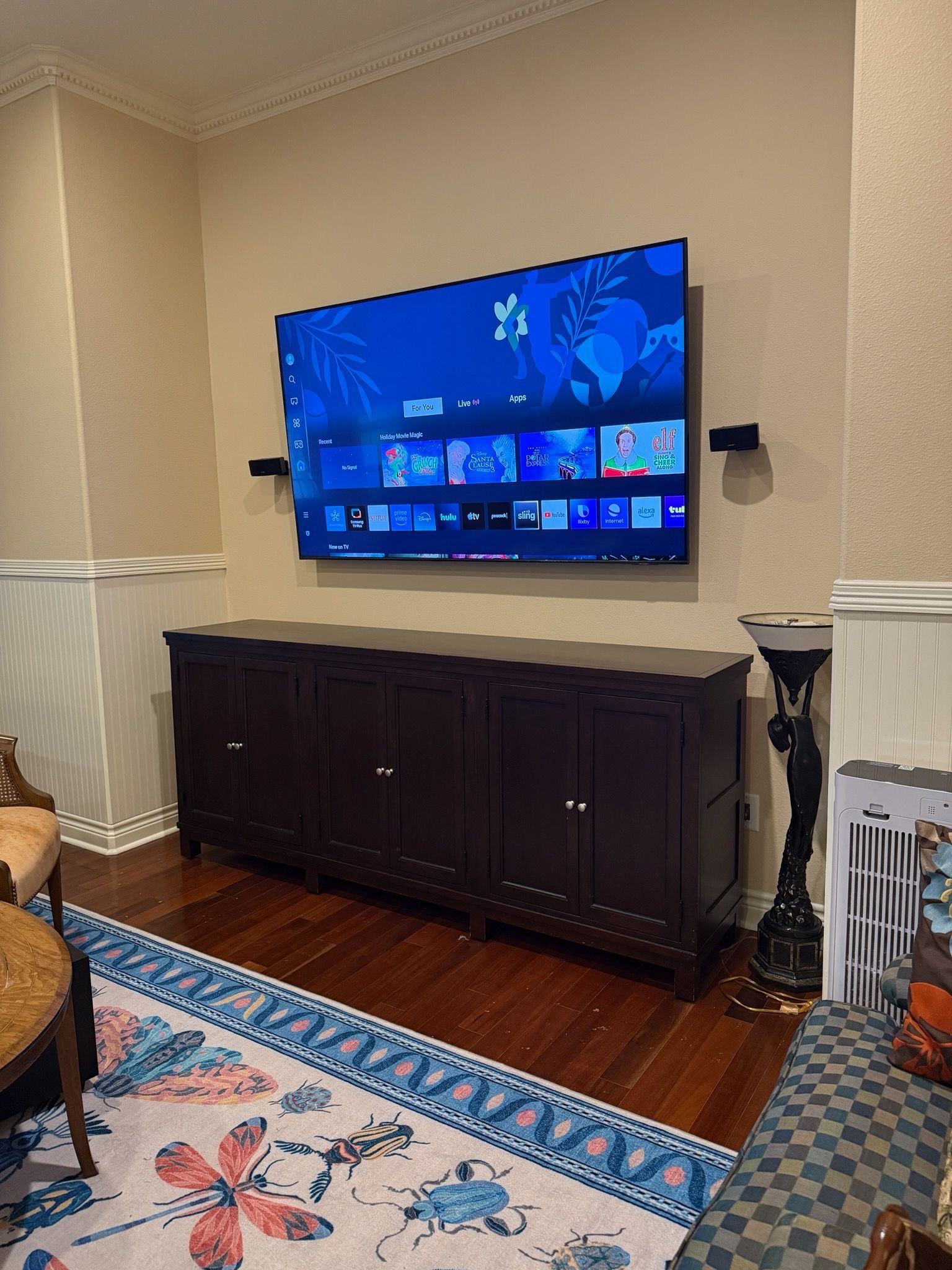 Television mounted above a dark brown cabinet against a beige wall, a patterned rug on the floor.