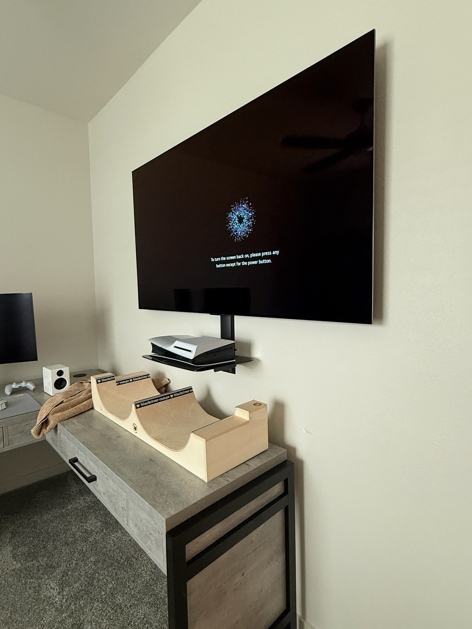 Wall-mounted TV with black screen above a desk. A media shelf and a ramp structure sit below.