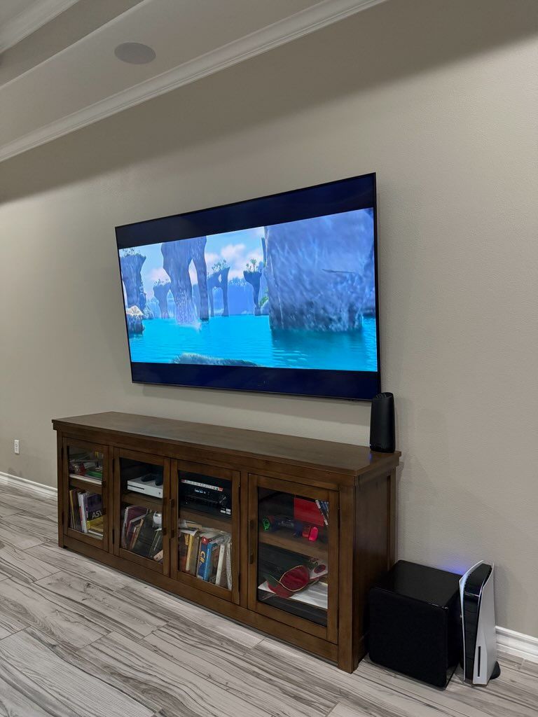Large TV mounted above a wooden media cabinet, a black speaker, and a game console against a beige wall.