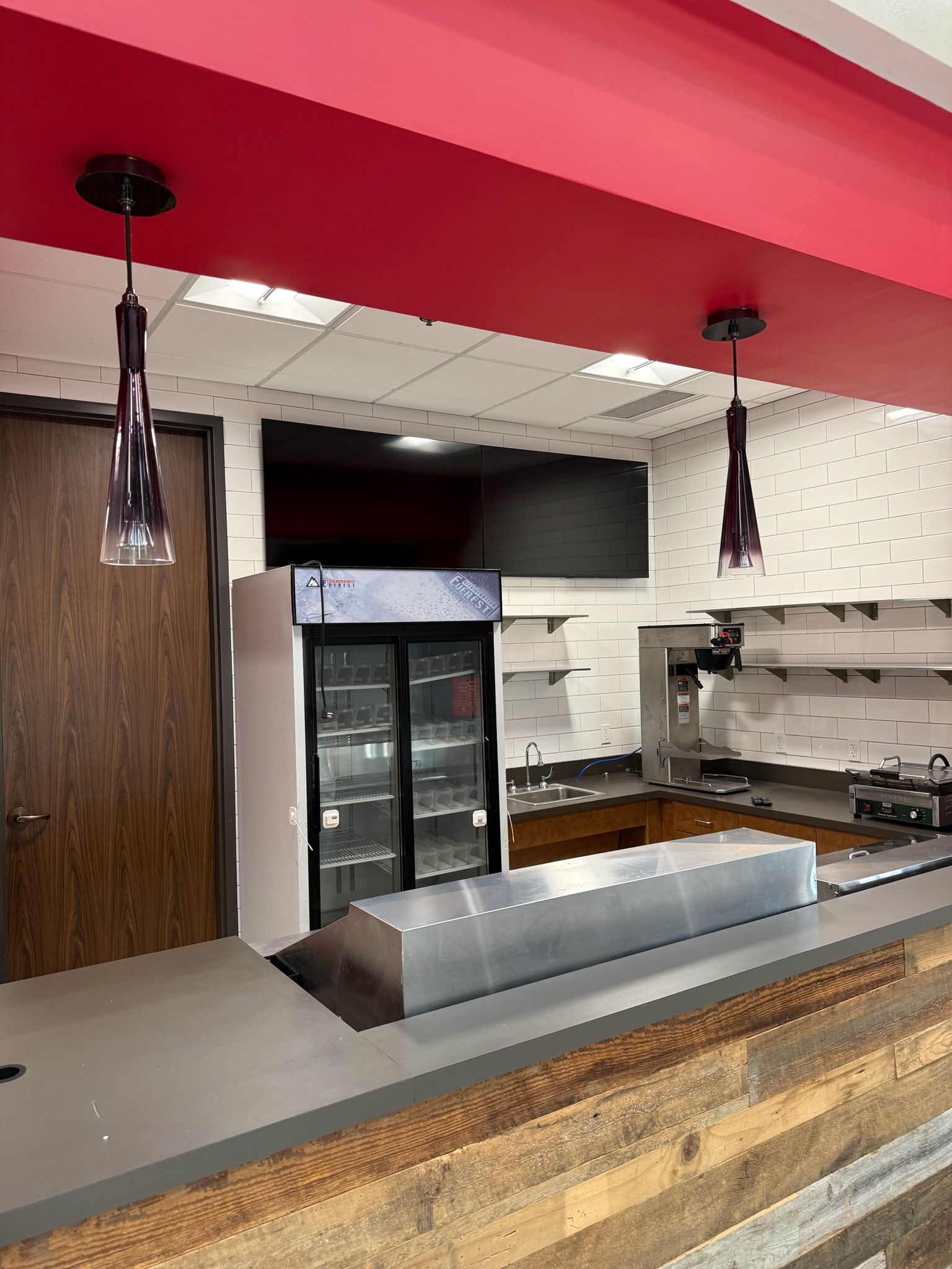A small commercial kitchen counter with a glass-front refrigerator, metal workspace, and two hanging purple pendant lights.
