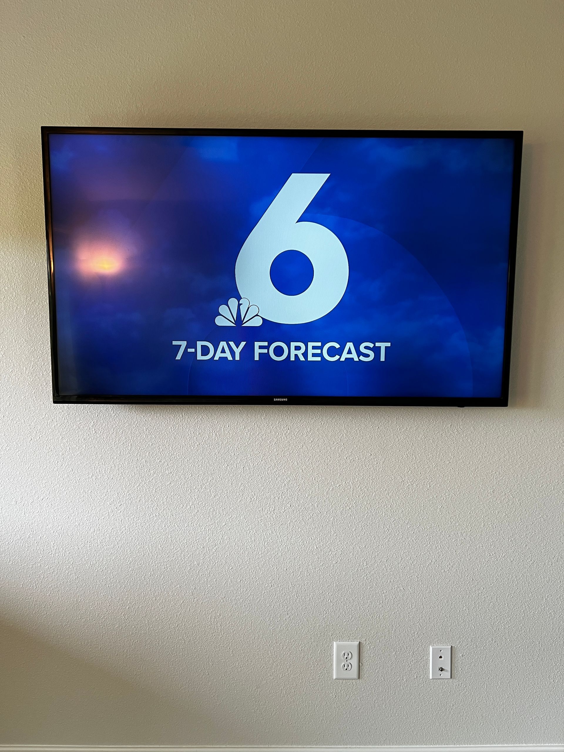 A television is hanging on a wall and shows a 7 day forecast