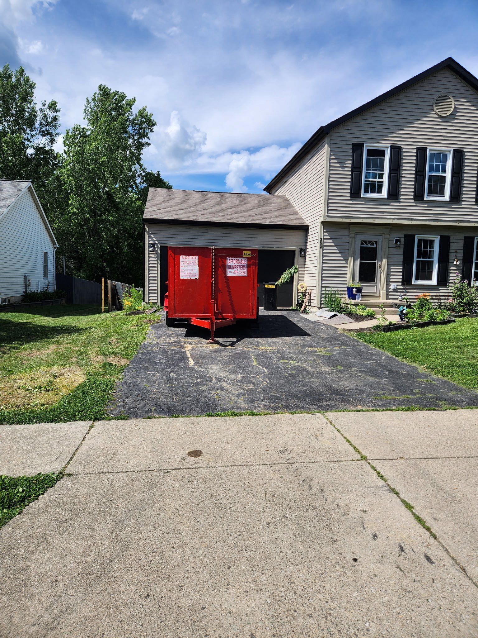 Red dumpster on a driveway beside a gray house and garage under a sunny sky