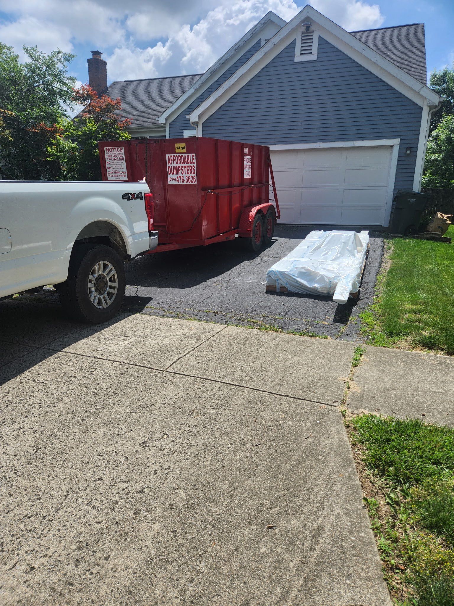 White pickup and red dumpster in a driveway beside a gray house; rolled roofing materials on the ground.