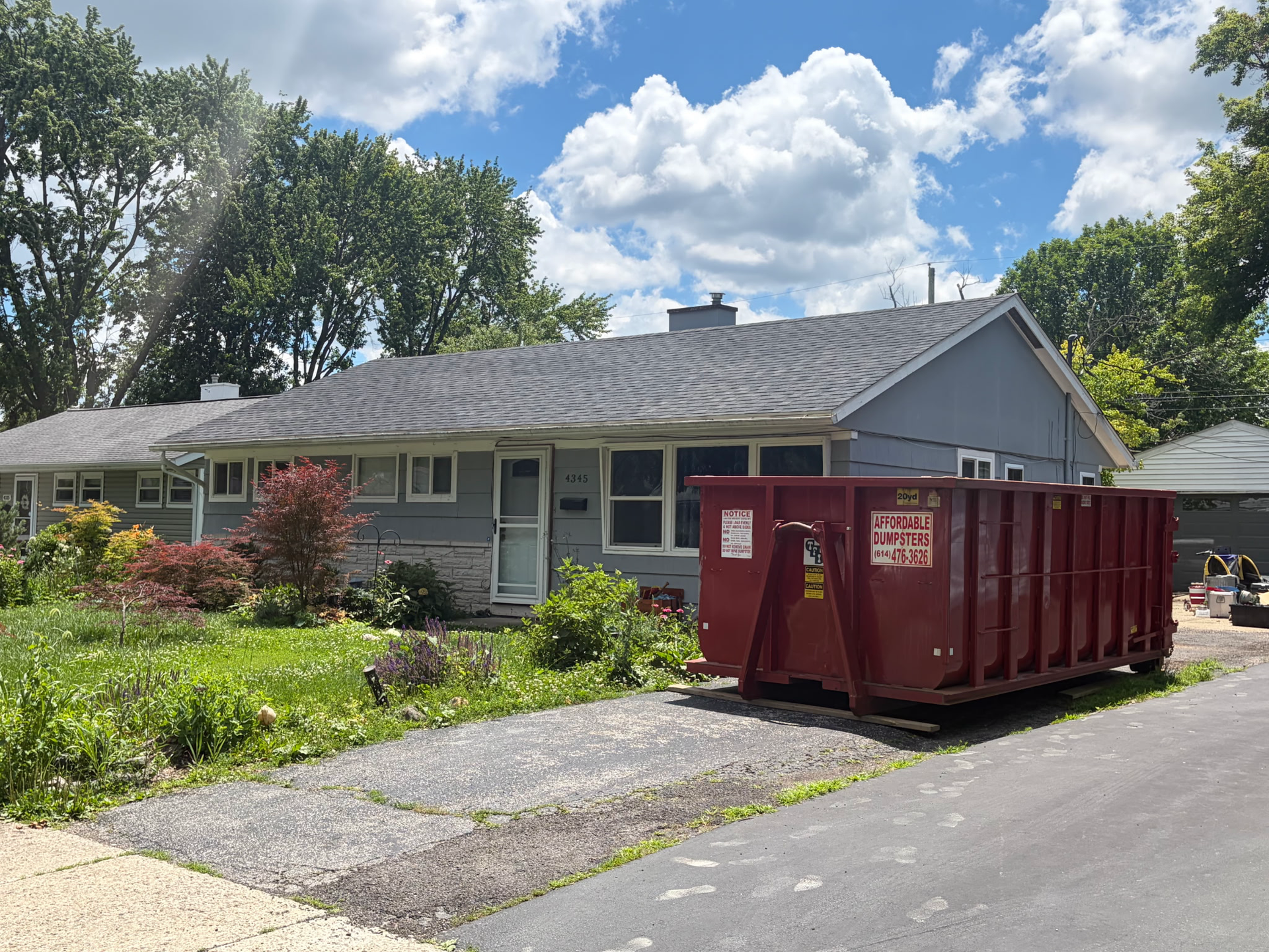 Gray house with a red dumpster in the driveway, surrounded by greenery under a cloudy sky Gray house with a red dumpster in the driveway, surrounded by greenery under a cloudy sky