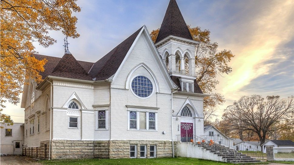 White wooden church with a steeple and arched windows, set against an autumn sky with trees.