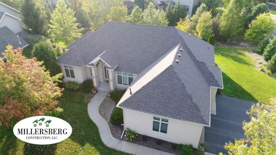Aerial view of a cream-colored house with a dark gray roof, lush green lawn, and surrounding trees.