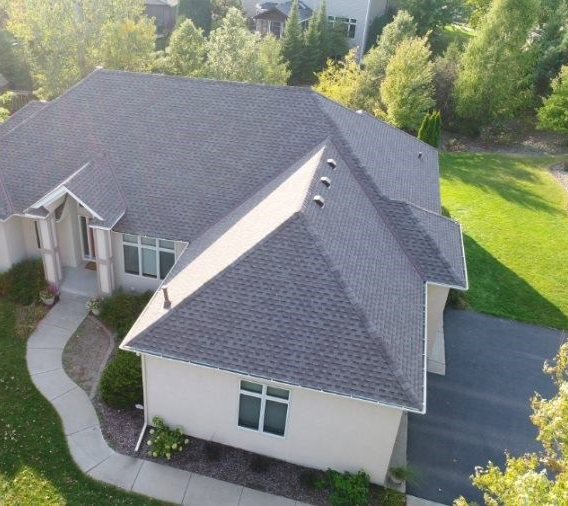 Overhead view of a house with a dark gray shingled roof, cream siding, and a curved walkway. Green lawn and trees surround the house.