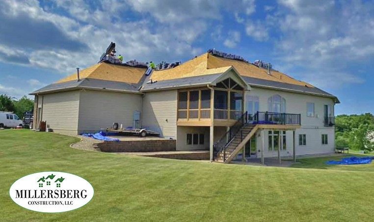 A large house with workers on the roof replacing shingles under a blue sky. Green grass in the yard.