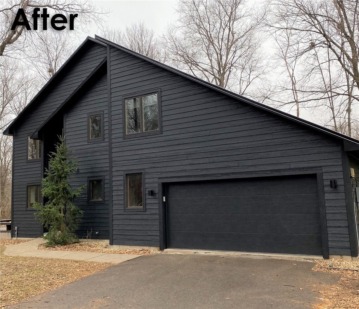 Black-sided house with a dark garage door, set in a yard with a driveway and trees.