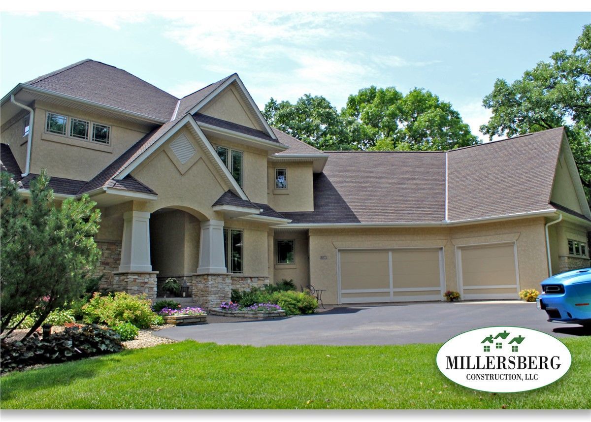 Beige two-story house with a three-car garage. Lush green lawn and mature trees.