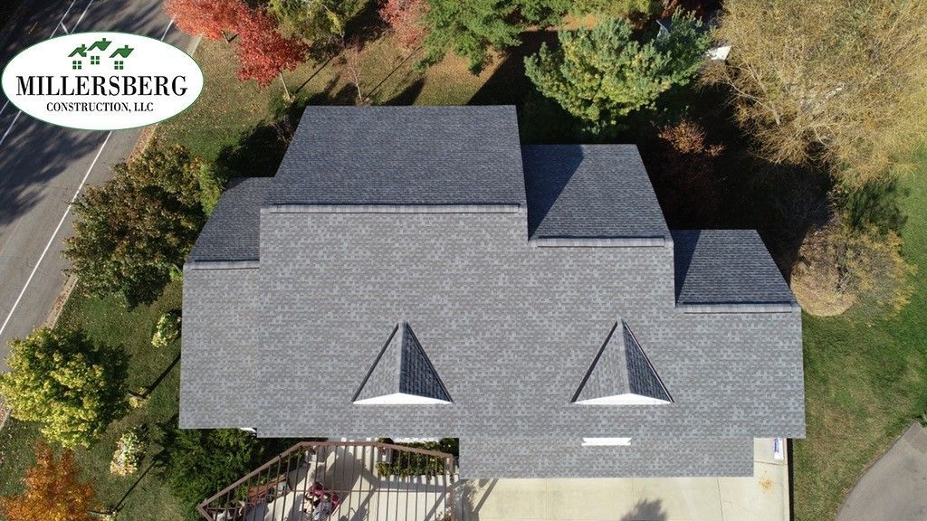 Aerial view of a gray shingled roof with a company logo in the corner. Autumn trees surround the house.