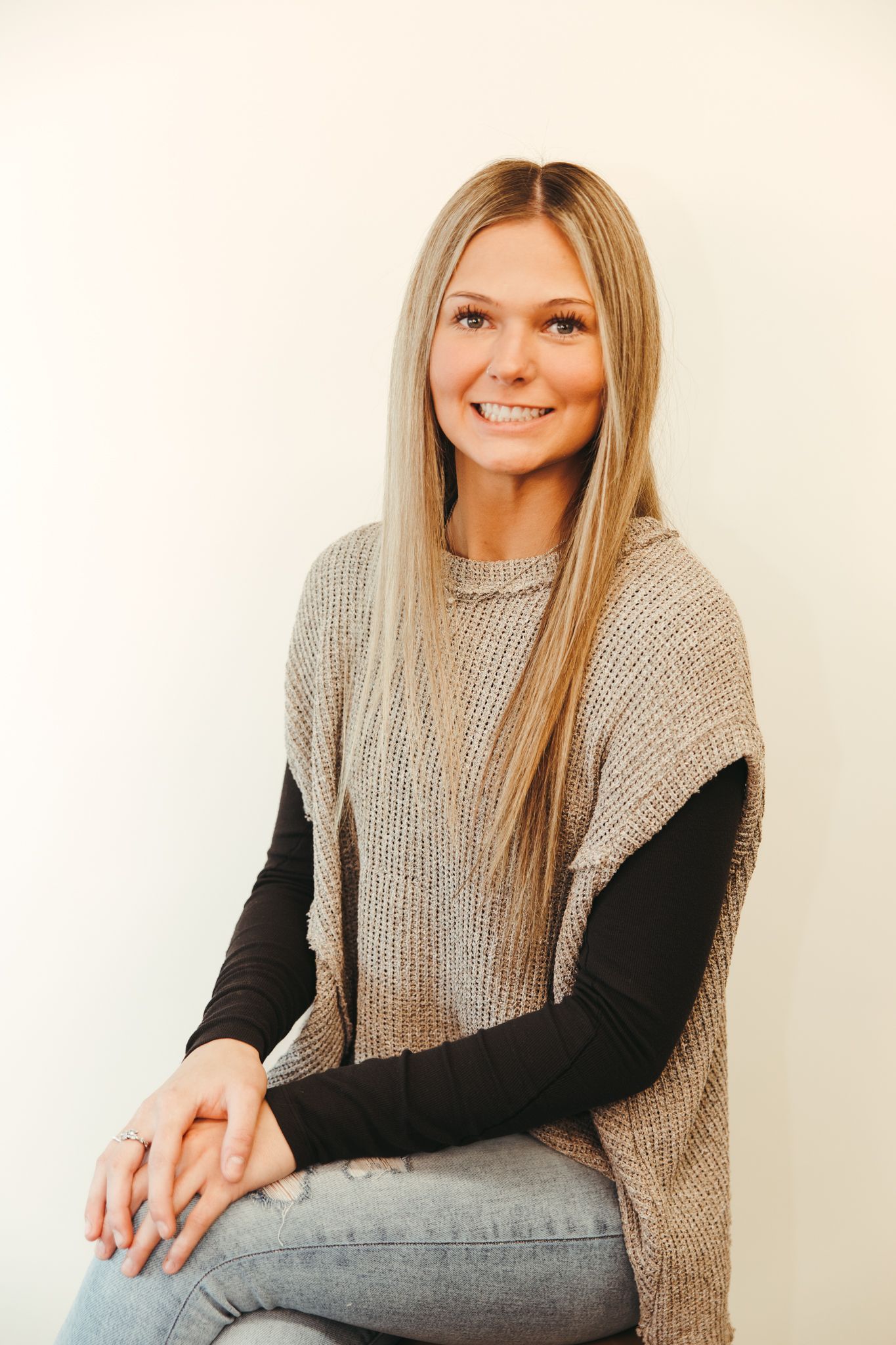 Woman with long blonde hair, smiling, wearing a gray knit vest, black long sleeve, and jeans.