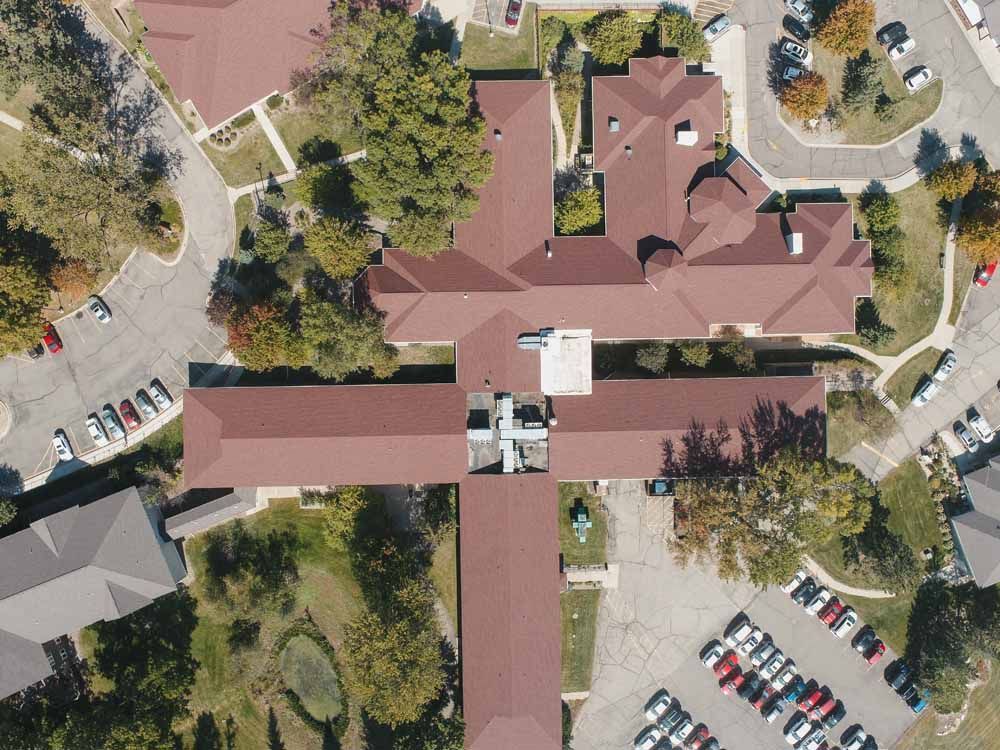 Aerial view of a complex with multiple wings, brown roofs, parking lots, and trees; likely a care facility.