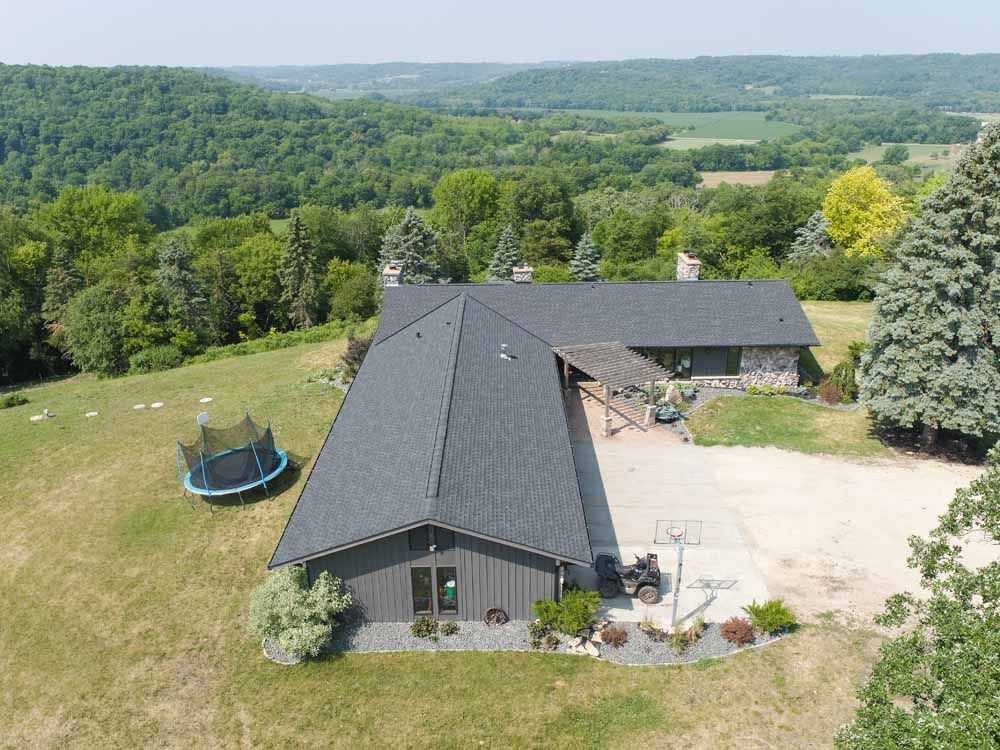 Aerial view of a gray-roofed house on a grassy hill, trampoline and forested background.