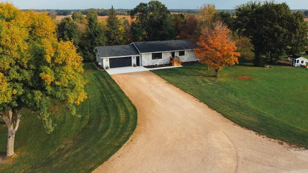 A one-story white house with a long gravel driveway, surrounded by green grass and colorful fall trees.