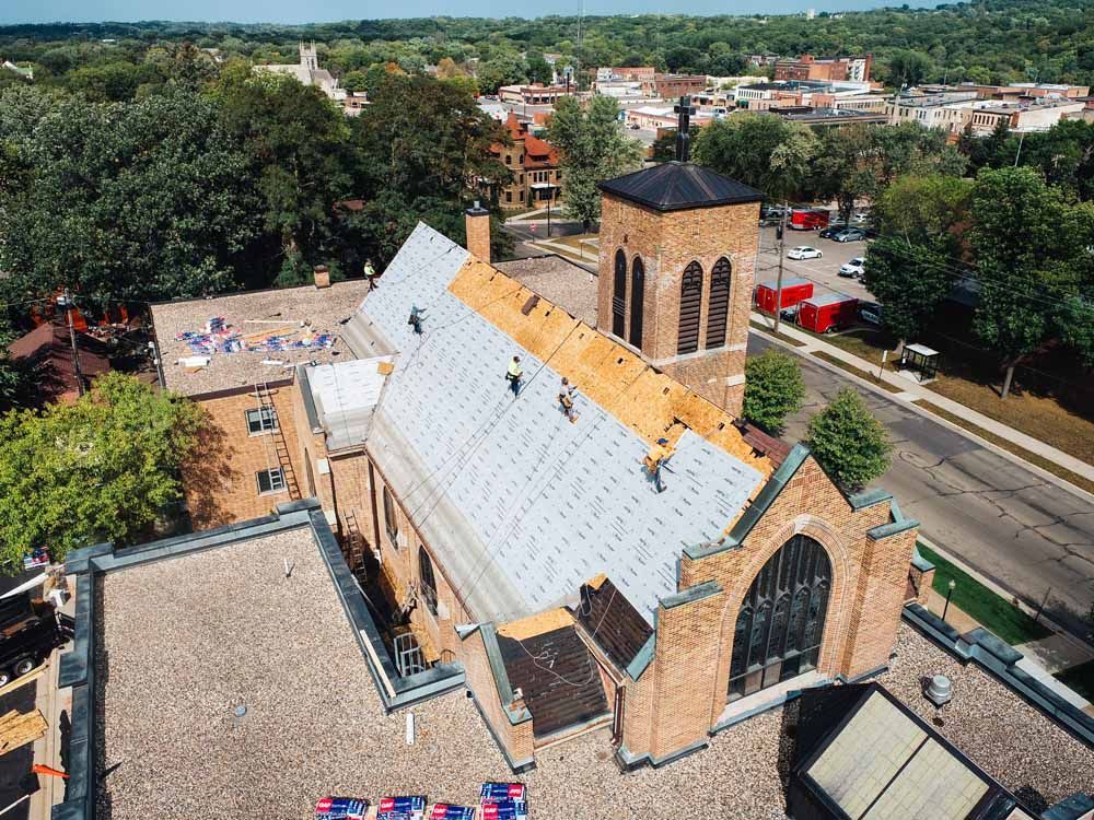 Church roof under construction; workers on rooftop, brick building, cityscape in background.