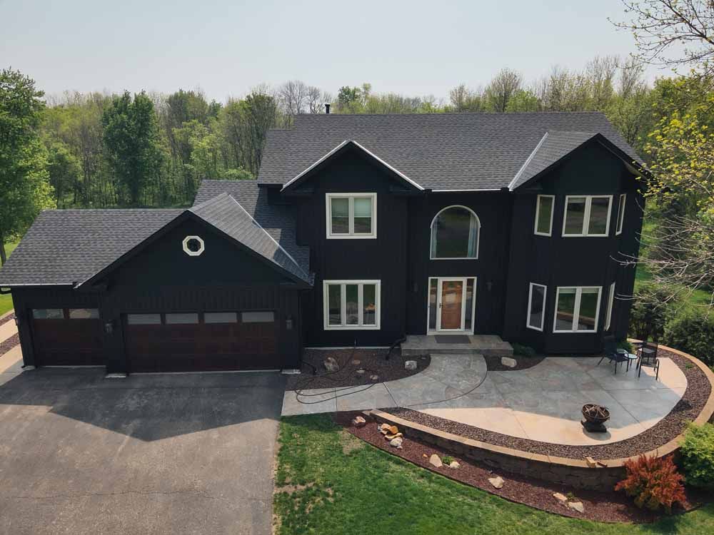 Black two-story house with a garage and a circular driveway surrounded by trees and a raised patio.