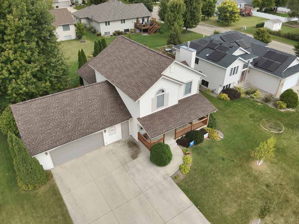 Aerial view of a two-story beige house with brown roof and attached garage, in a green lawn setting.