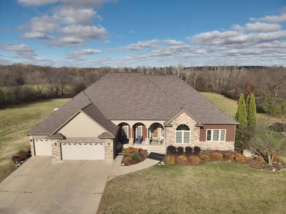 House with brown roof, stone and tan siding, garage doors, and front porch, set in grassy yard.
