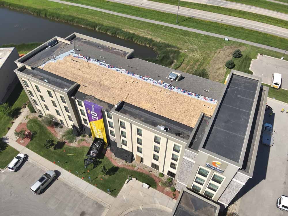 Aerial view of a hotel with a flat roof being repaired. Cars parked nearby. Green grass and a waterway are adjacent.