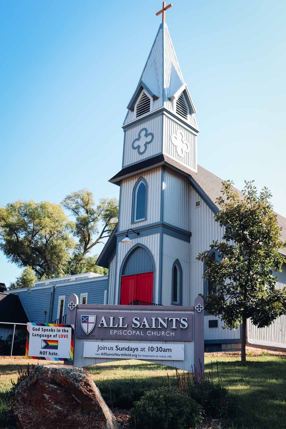 All Saints Anglican Church with steeple, cross, and sign. Red door, blue and white facade, and sign with service times.