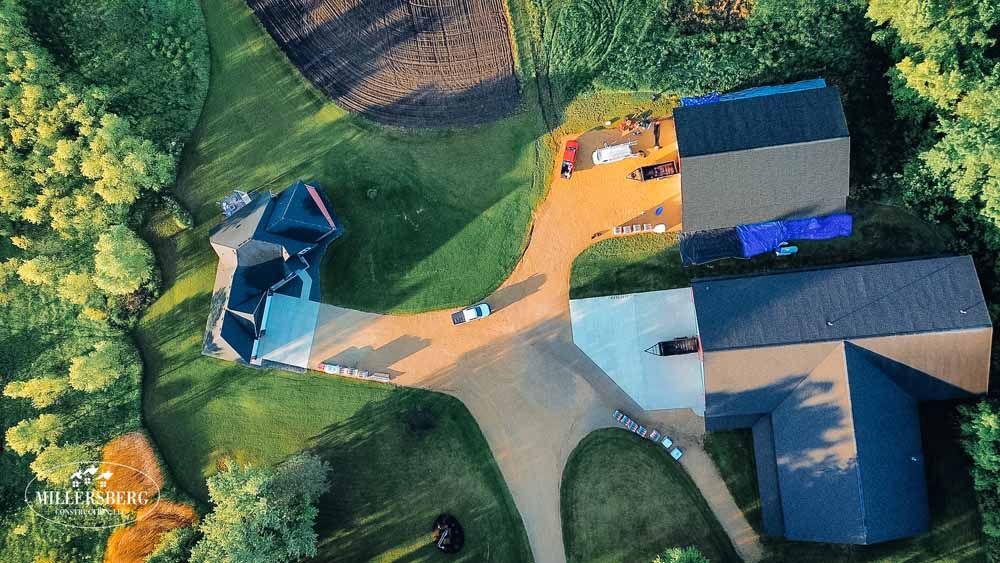 Aerial view of a house and outbuildings with driveway, surrounded by green grass and trees.