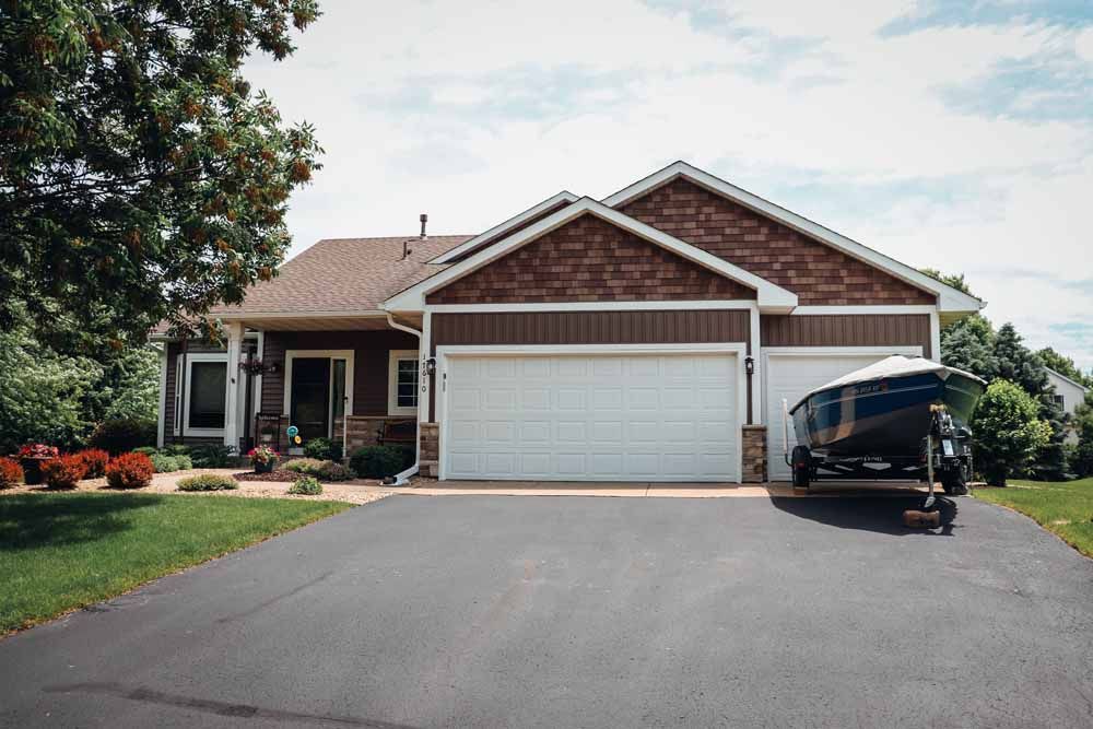 House with brown siding, white garage doors, and a trailer in the driveway on a sunny day.