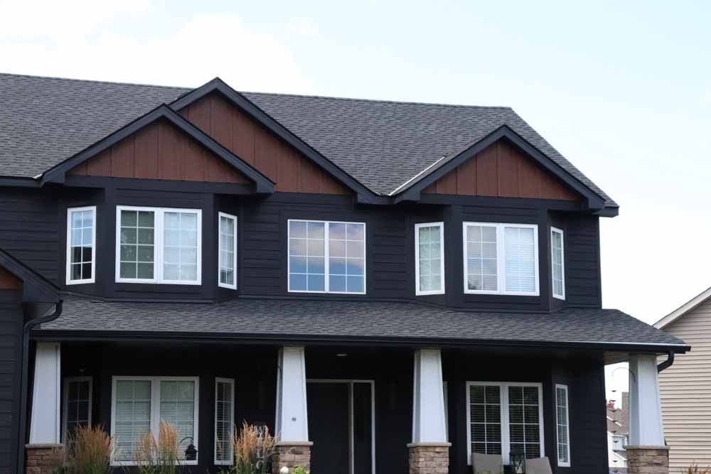 Black house with white-trimmed windows and brown accents on roof peaks; porch with white pillars.
