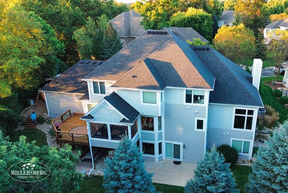 Large, two-story house with blue-gray siding, dark roof, surrounded by trees. A wooden deck and screened porch are visible.