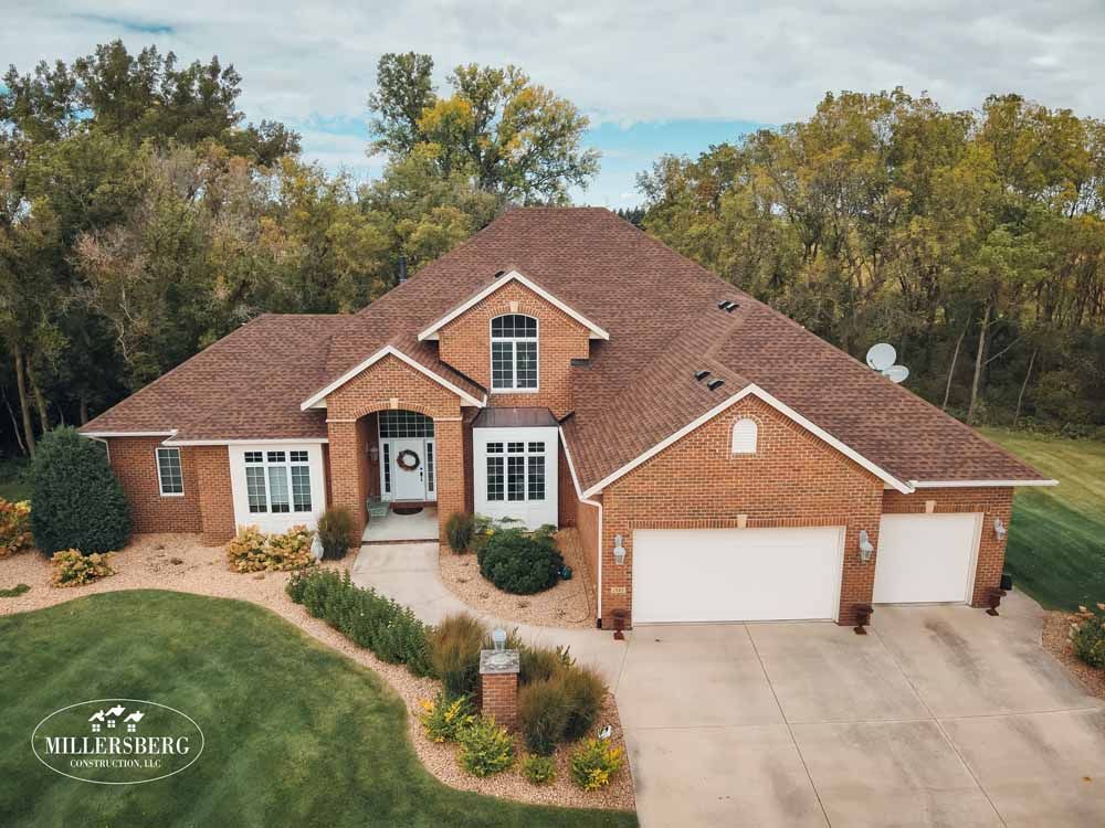 Brick house with brown roof, three-car garage, and landscaped yard. Trees in background, cloudy sky.