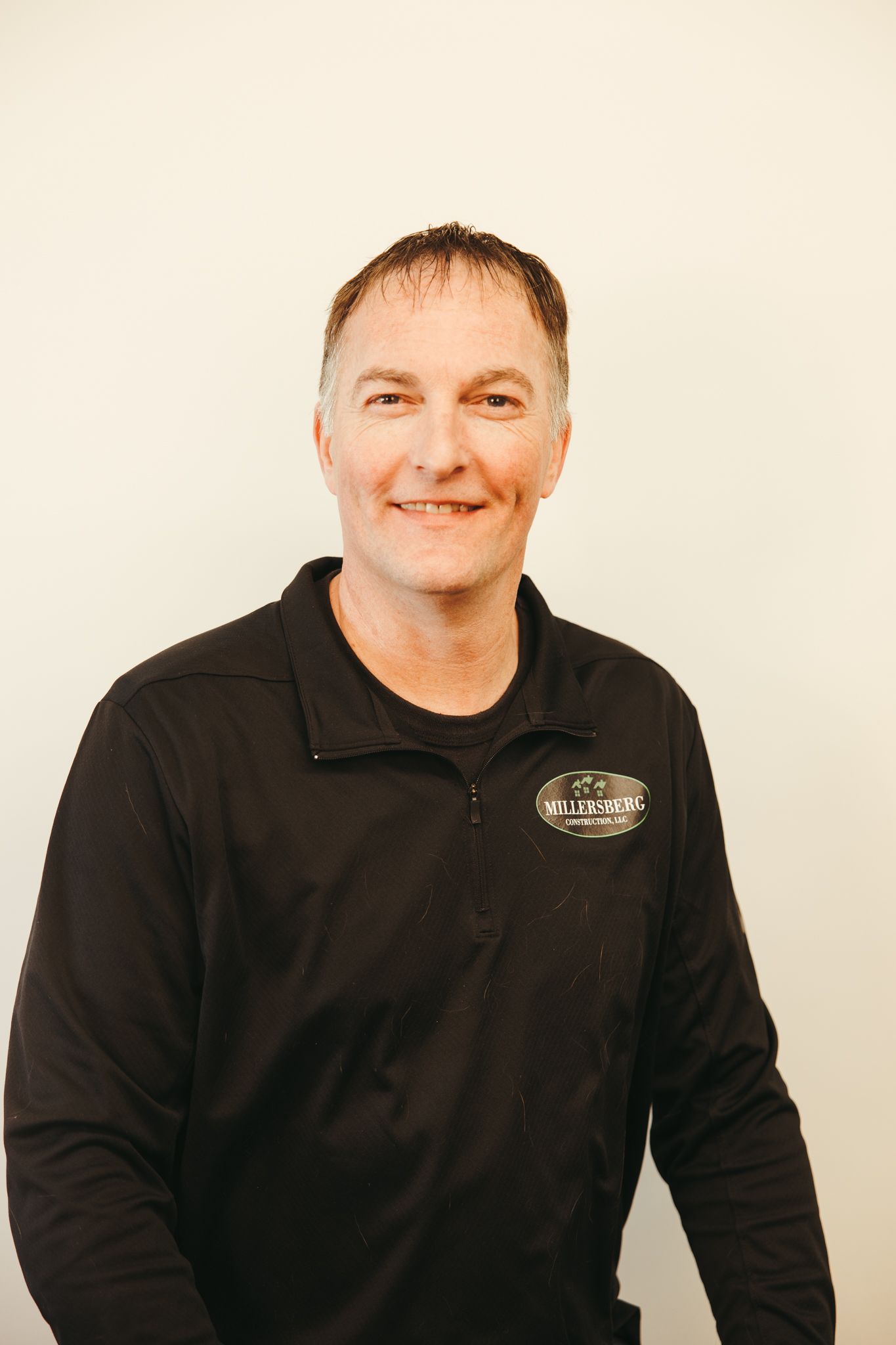 Man in black shirt smiling at the camera, standing in front of a white wall.