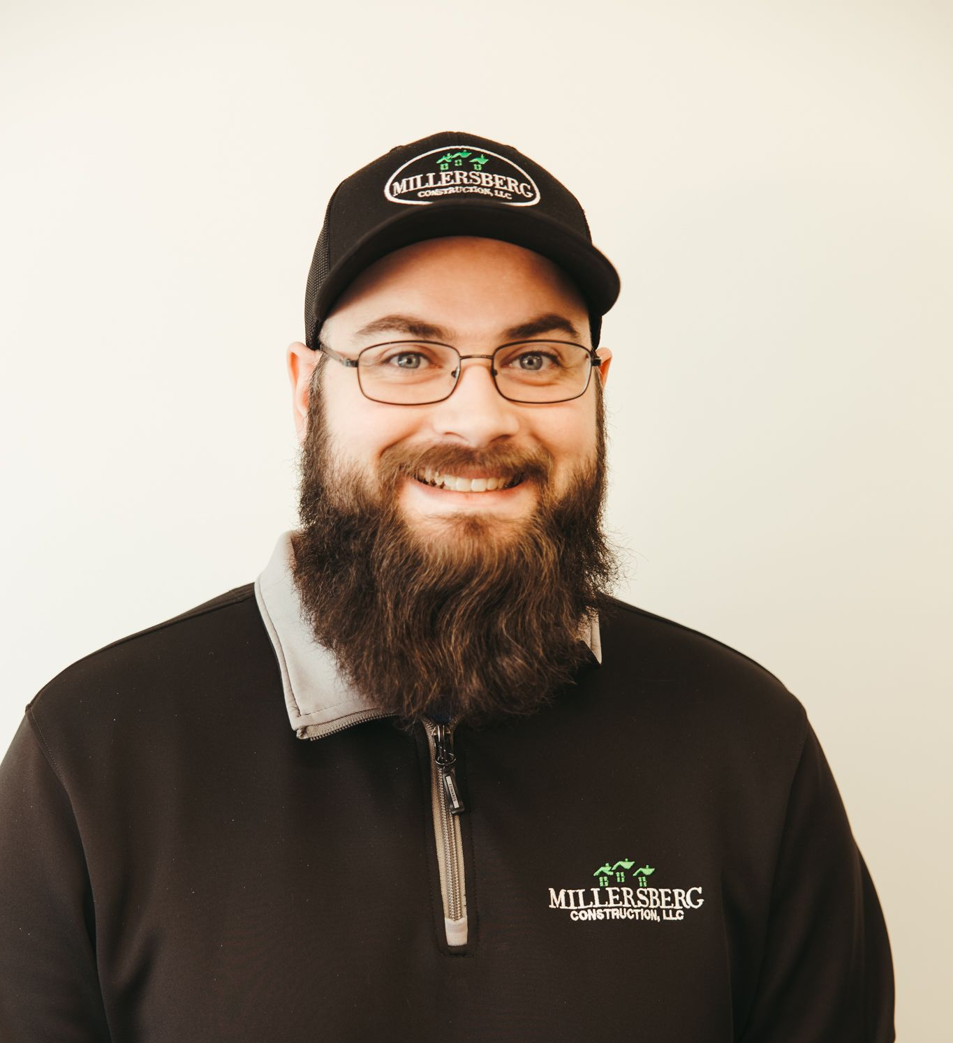 Man with glasses and beard, wearing a hat and black sweater with a logo, smiling in front of a white wall.