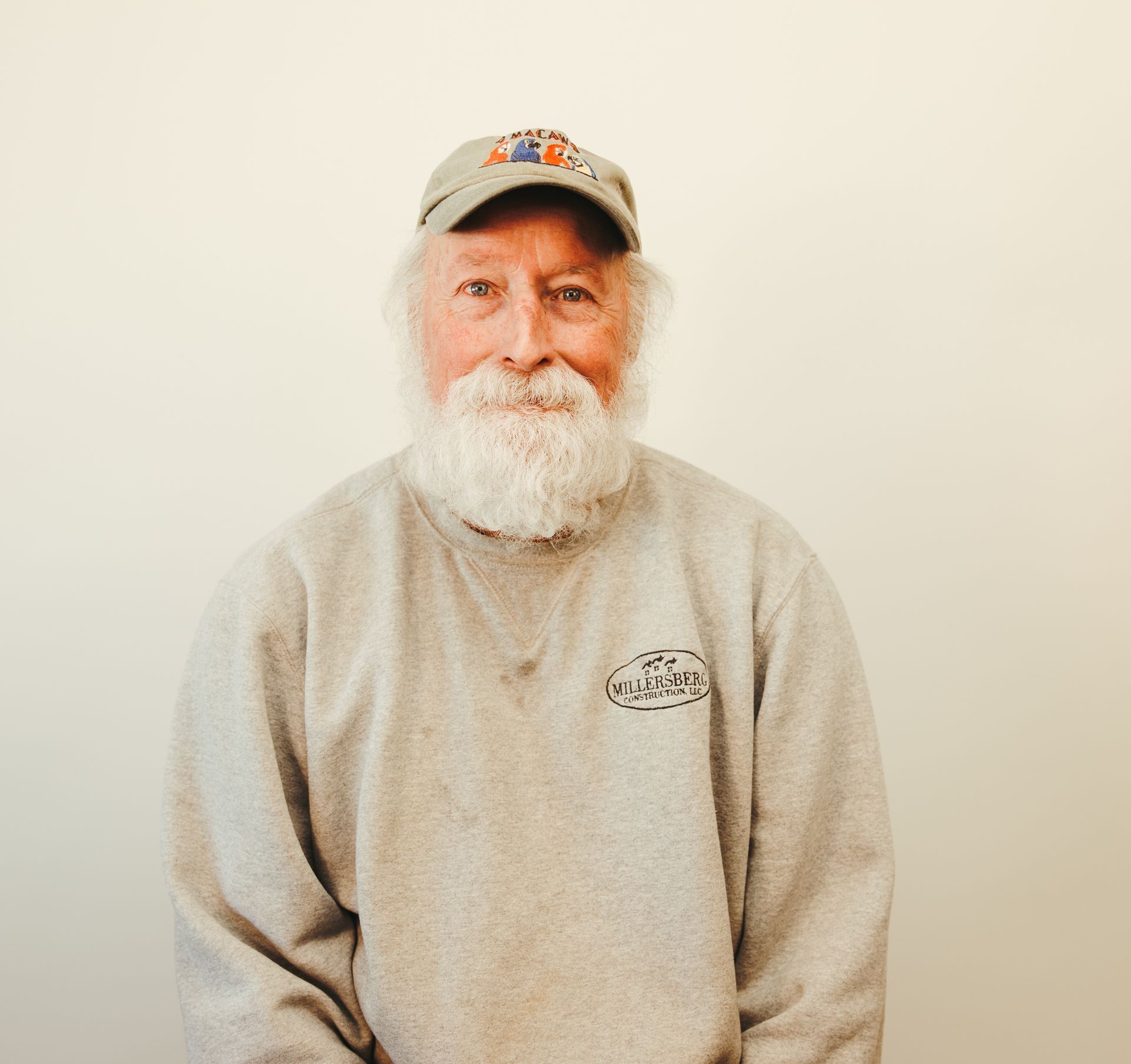 Man with a long white beard and hat, wearing a gray sweatshirt, smiles.