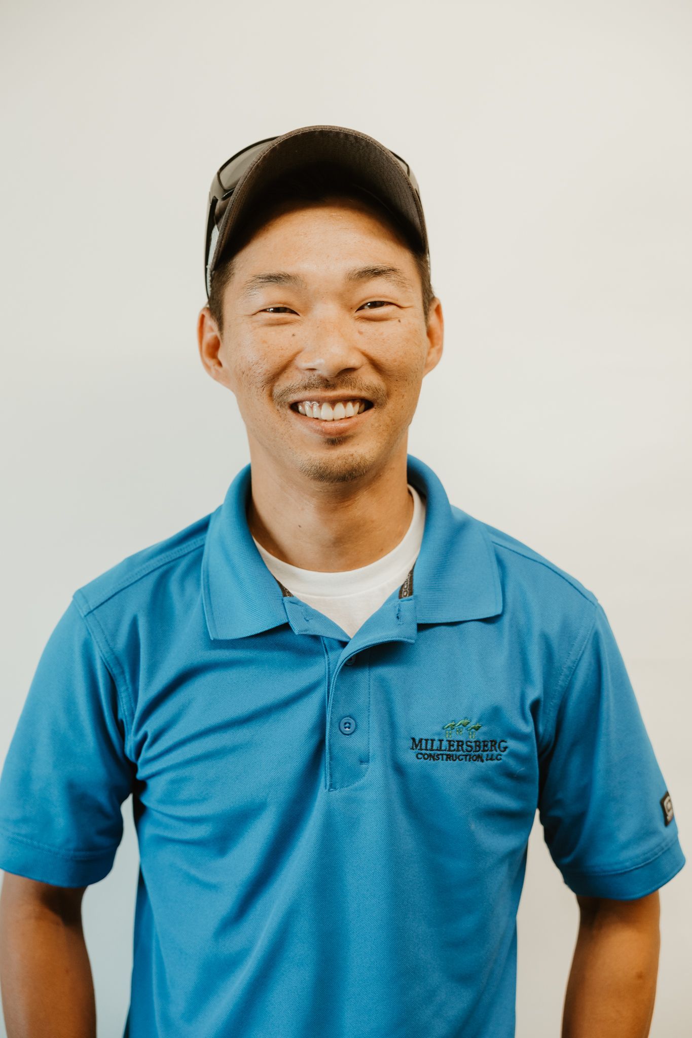 Man smiling, wearing a blue polo shirt and a cap, standing in front of a white wall.