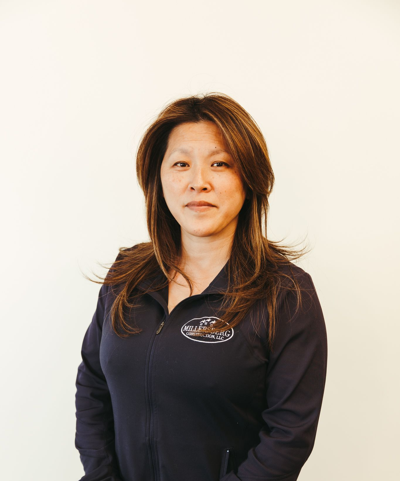 Woman with long brown hair wearing a dark blue zip-up jacket smiles in front of a white wall.