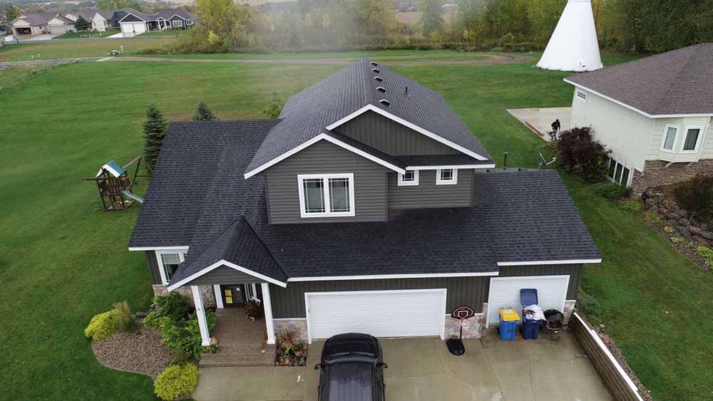 Dark gray house with a black roof, white trim, and a car in the driveway on a grassy lot.