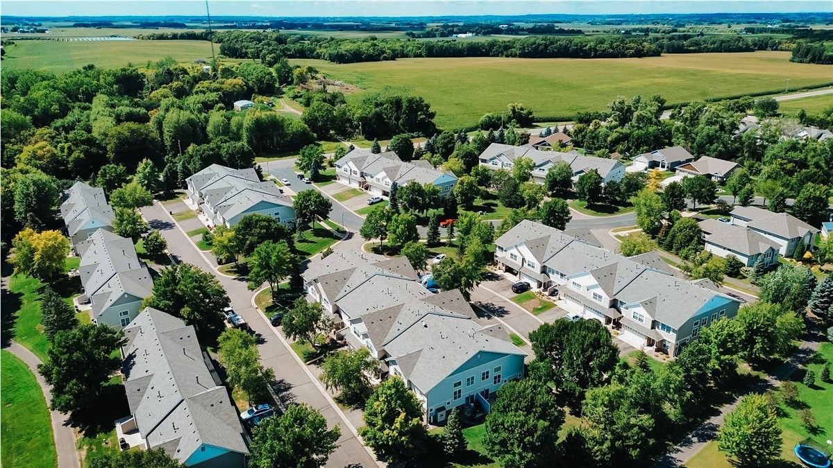 Aerial view of a suburban neighborhood with gray-roofed houses surrounded by green trees and fields.