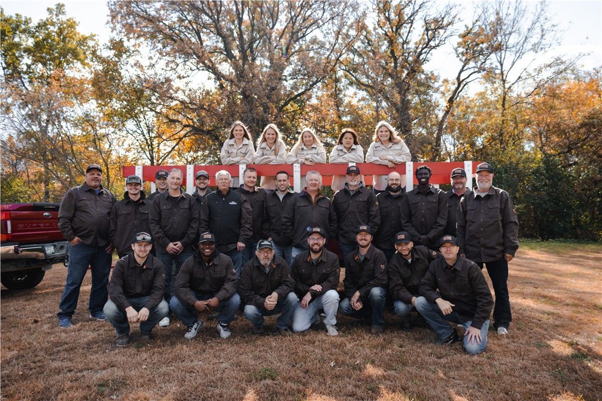 Group of people posing outdoors; men and women in black jackets, red and white backdrop, fall setting.