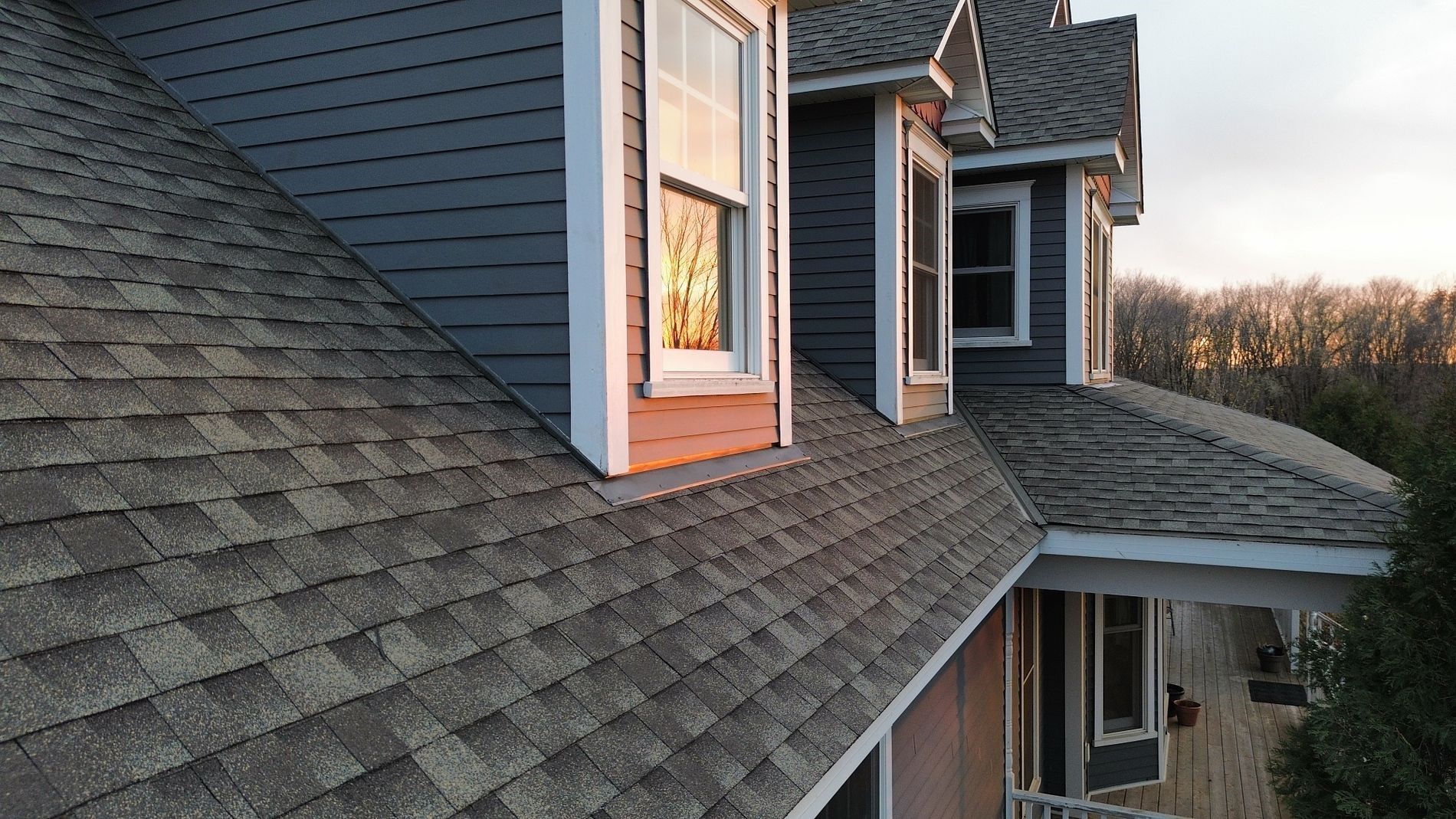 Gray shingled roof of a house with dormer windows and blue siding at dusk.