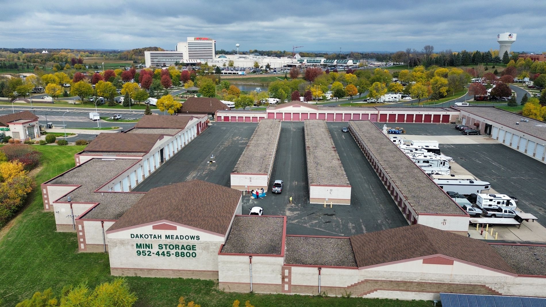 Aerial view of B & M Storage facility. Rows of storage units with brown roofs, large paved area, and surrounding trees.